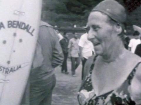 Female surfer from the 1960s and 1960s 'Ma' Bendall, standing next to a surfboard on the beach, she is pictured from the chest up and wearing a swimsuit and swimming cap.