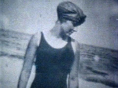 Waist up image of a young woman at the beach, dated roughly from the 1920s or 1930s. She is wearing a black swimsuit and has a swim cap over her hair, she looking to the side and down towards the sand. 