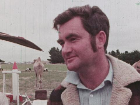 Man in a paddock wearing a lumber jacket. A camel behind him.