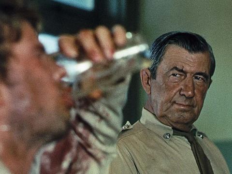 A country police officer played by Chips Rafferty watches while John Grant drinking a glass of beer