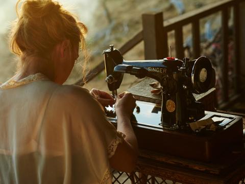 Kate Winslet in a scene from The Dressmaker. She is seated in front of a vintage sewing machine with her back to the camera and her hands are visible, threading cotton through the machine.