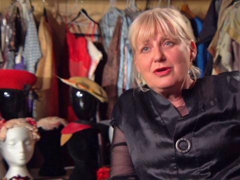Head and shoulders shot of costume designer Marion Boyce looking directly at camera. She is wearing a black blouse. There are dresses hanging on racks, along with wigs and hats on mannequin heads in the background.