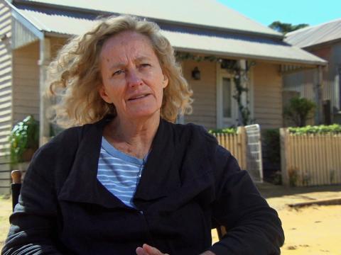 Margot Wilson, costume designer, pictured from the waist up wearing a striped shirt under a cardigan sitting outdoors in front of an old house that is part of the film set for The Dressmaker