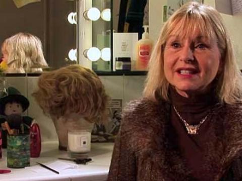 Actress Liza Goddard sits in front of a dressing room mirror, with a wig and make-up jar on the tabletop next to her