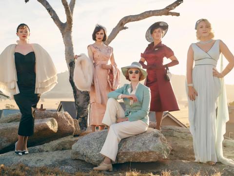 Five of the women from the cast of The Dressmaker, standing on a rock formation in the Australian outback, posing for the camera wearing 1950s high end fashion pieces.