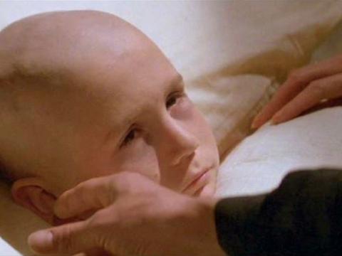 A pair of hands hold the face of a sickly, bald child lying in bed.