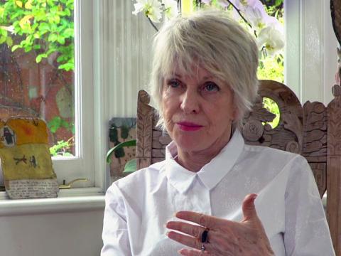 Head and shoulders shot of film editor Jill Bilcock talking to camera and seated on an ornately carved wooden chair in front of a window with trees visible outside.