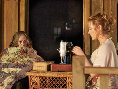 A scene from the film The Dressmaker showing Judy Davis sitting in a chair with a blanket pulled up to her nose and looking sideways at Kate Winslet who is seated in profile in front of a vintage sewing machine.
