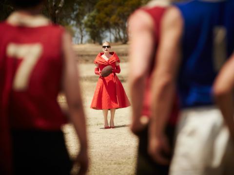 Full length shot of actress Kate Winslet in a striking red dress and sunglasses, holding a football. In the foreground, slightly out of focus are a group of football players with their backs to the camera all looking at Winslet. 