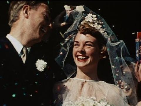 A bride dressed in a white dress and veil smiles at her groom.