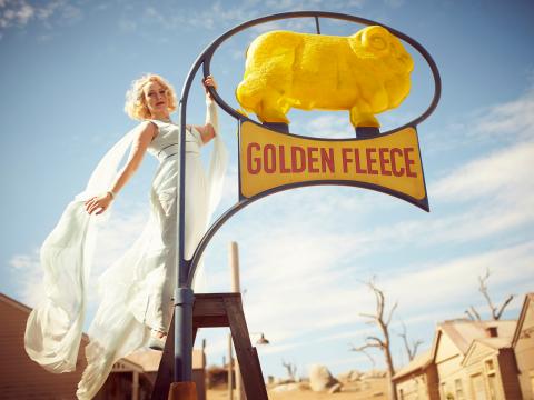 Actress Rebecca Gibney wearing a flowing, powder blue tailored gown is on a ladder holding onto a street light/sign that says 'Golden Fleece' with a sheep above it, in a scene from the film The Dressmaker.