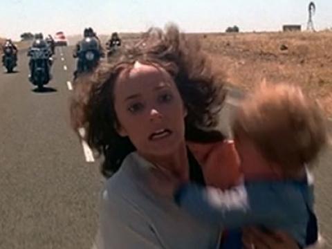 Woman runs down a road in terror holding a child while a motorcycle gang chases her.