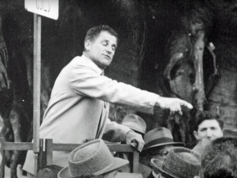 A middle-aged man addresses a group of people in a park.