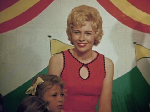 A young woman smiling directly at a camera in front of a brightly coloured backdrop. A small child is seated in front of her.