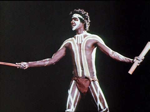 David Gulpilil dancing holding wooden sticks. He is wearing body paint on his face and body.