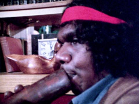David Gulpilil playing didgeridoo with a red headband around his head.