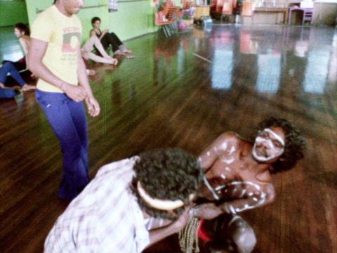 David Gulpilil in full body paint dancing with another man in a hall in Redfern.