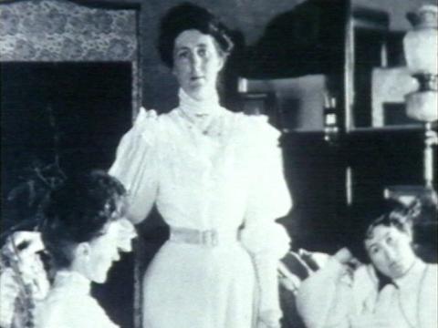 A woman standing with two women seated beside her, black and white, dressed in clothing of late 1800s