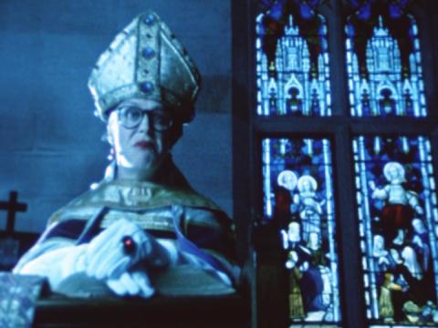 A woman dressed as a stern 16th century bishop standing beside stained glass windows 