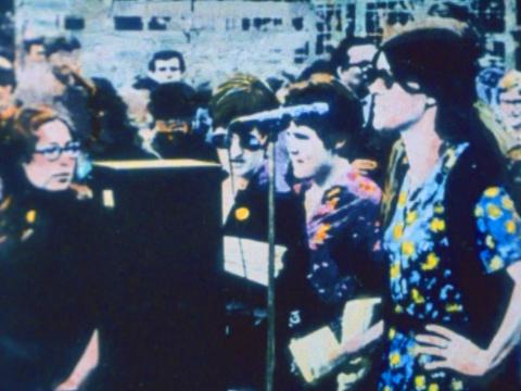 Colourised black and white image of women standing at a rally with a microphone in frame.