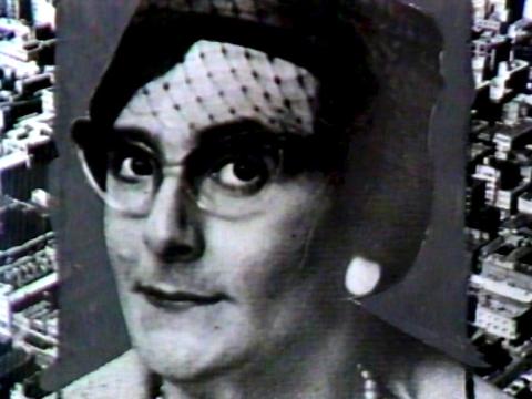 Edna Everage (Barry Humphries) in a black and white photograph from early in her career. She has a somewhat accusatory look. The photo is on top of a background of suburbia from the air.