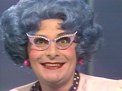 Dame Edna Everage circa 1975 with her wisteria bouffant, winged spectacles and red lipstick. She looks directly at the camera.