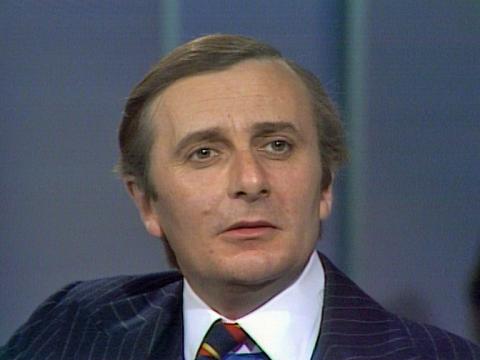 Comedian Barry Humphries in close-up. He's dressed in a suit and tie.