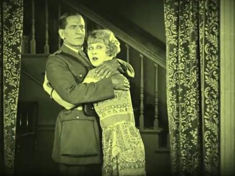 A couple embrace in front of a staircase with curtains on either side. They both have concerned looks on their faces. The image is from a scene in the silent era film Empire Builders (1924).