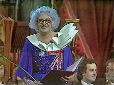 Dame Edna Everage is wearing a dress with an Australian flag made out of sequins and a collar in the shape of the Sydney Opera House.