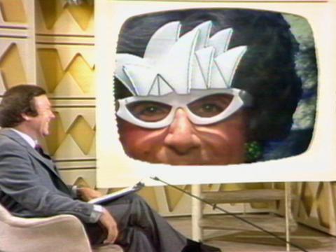 Dame Edna Everage in close-up wearing her Sydney Opera House insired spectacles. She appears inset on a screen that features Mike Walsh interviewing her.