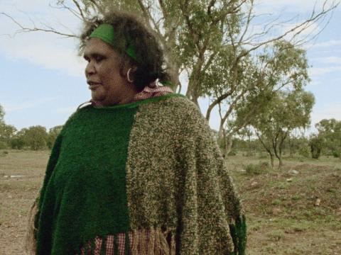 Aboriginal director, Essie Coffey, standing in a scrubland area, looking off camera. She is wearing a knitted shawl and a headband and earrings.