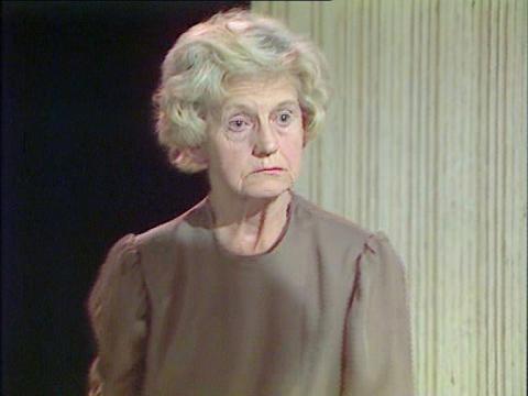 Dame Edna's Everage's bridesmaid, Madge Allsop, played by Emily Perry looks blankly off into space in a grey smock dress.