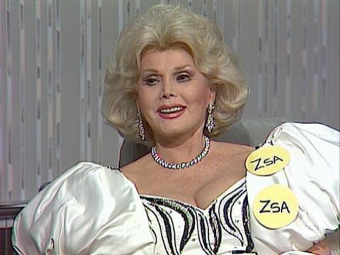 Actor Zsa Zsa Gabor is sitting in a studio being interviewed. She is wearing two badges, each saying 'Zsa' on her puffy white dress bodice.