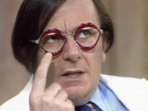 Comedian Barry Humphries in close-up. He's pointing to the pair of spectacles he's wearing with lips and teeth on them. They obviously belong to Dame Edna Everage.