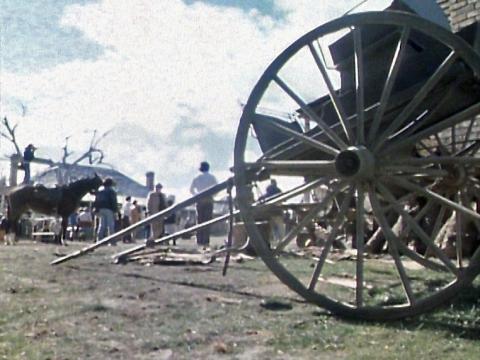 Close up view of a wagon wheel attached the back end of a wagon. There are a group of men and horses standing around in the background. 