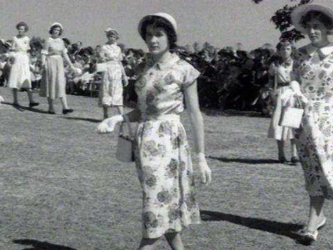 A woman is walking in a parade with other women. They wear dresses, hats and gloves.