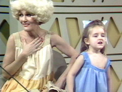 Jeanne Little has her hand to her heart and is looking off-screen. A young child is singing next to her.