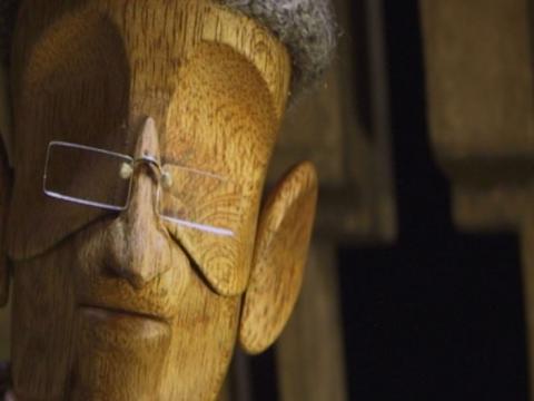 Animation still of a wooden face wearing glasses but has no eyes.