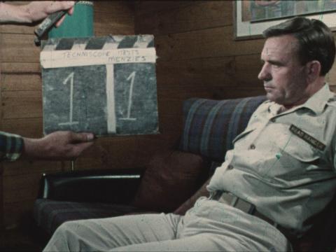 Ed Devereaux slouches on a couch with a crew member about work a clapper board.