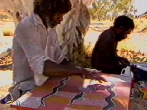 Two indigenous artists seated beneath a tree painting.
