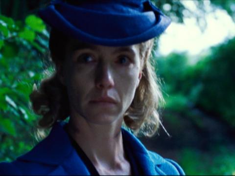 Actor Ursina Lardi is standing outside looking at someone off-screen with tears in her eyes. She is wearing a striking blue hat and jacket.