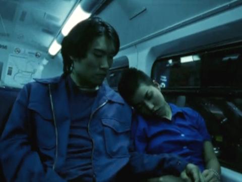 A man and a woman are on a train at night. The woman is sleeping and the man is about to touch her hand.