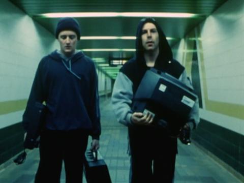 Two men walk inside a tunnel at a railway station. One of them is holding a television.