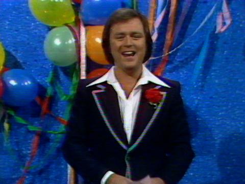 Young Talent Time host Johnny Young is wearing a shirt and suit jacket. There are balloons and streamers in the background.