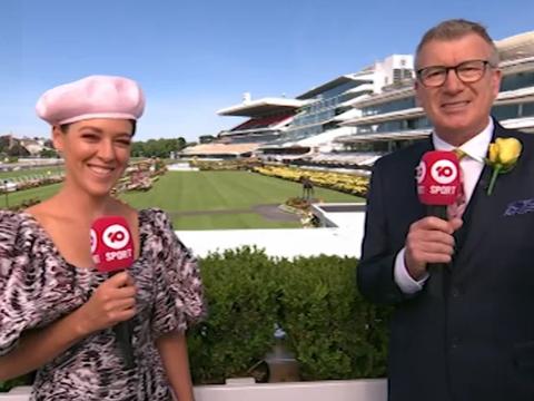 Two television presenters holding microphones and smiling at camera.  They are dressed in formal wear and standing in front of a racecourse.