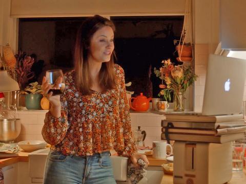 A woman stands in a kitchen holding a glass of wine and looking at her laptop computer.
