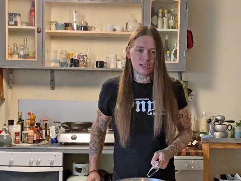 A man with long brown hair and tattooed arms stands in a kitchen, looking directly at the camera. 