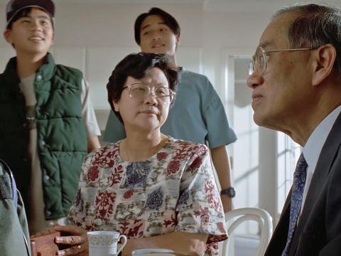 A middle-aged Asian couple sit at a kitchen table. Two teenage Asian boys stand behind them smiling.