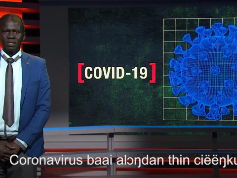 A newsreader stands in front of a big screen that says COVID-19 with a symbol representing the virus. At the bottom of screen are subtitles in the Dinka language