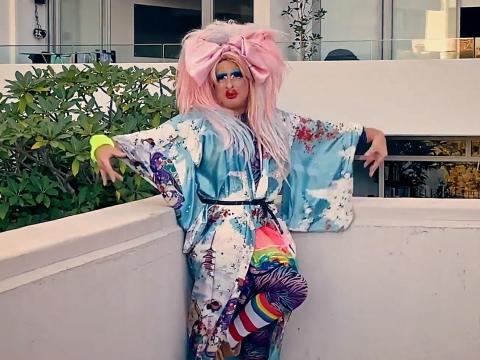 A drag queen with outrageous, bright makeup and big pink hair poses on a rooftop with arms outstretched.